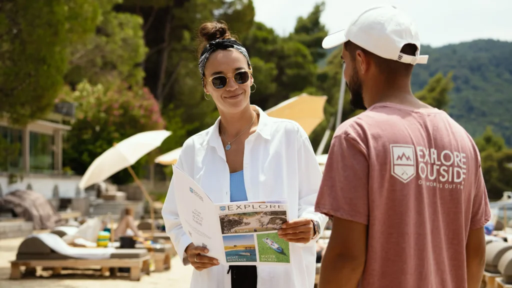 Hotel receptionist using a printed price list and digital cruise guide to assist guests at the front desk